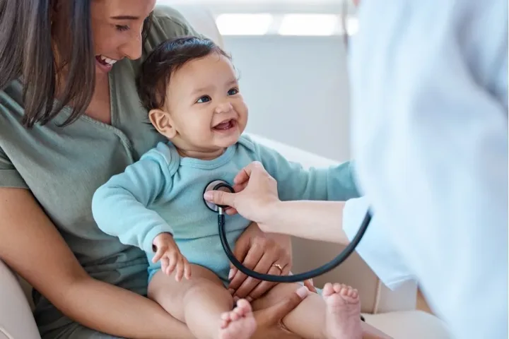 A doctor listening to a smiling baby’s chest while a parent holds them